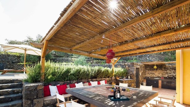 An outdoor dining area with a stone wall, wooden chairs, a table with a glass top, and a thatched roof.