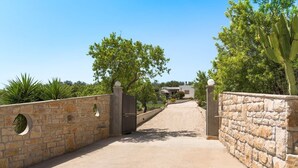 A paved driveway leading to a gated entrance with stone walls and a modern building in the background. - Villa Raggera - Private villa with pool near Noto (Ispica)