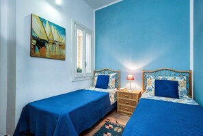 Ein Zimmer mit zwei Betten, jeweils mit blauer Bettdecke und passenden Kissen, einem Holz-Kopfteil, einem Weiden Nachttisch mit Lampe und einem Gemälde an der Wand.