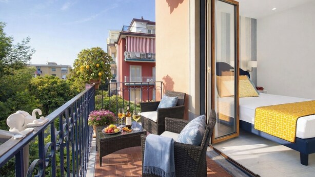 Een balkon met rieten meubels, een bed met een geel sprei en uitzicht op omliggende gebouwen.