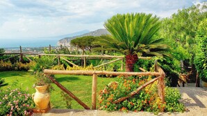 Un jardín con cerca de madera, una palmera y vista al mar y montañas al fondo.