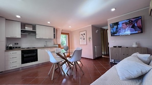 Une cuisine moderne avec des meubles blancs, un coin repas doté d’une table et de chaises en verre, et un téléviseur fixé au mur.