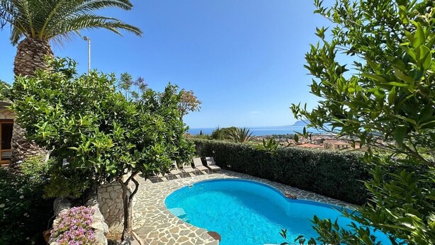 Une piscine à bord incurvé, entourée de carreaux de pierre et d’une végétation luxuriante, offrant une vue sur un paysage côtier.