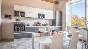 Una cocina moderna con armarios blancos, horno empotrado y comedor con mesa de vidrio y sillas blancas.