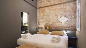 A modern hotel room with a large bed, two pillows, a small table, and a brick wall. - Porxos Garden (Barcelona)