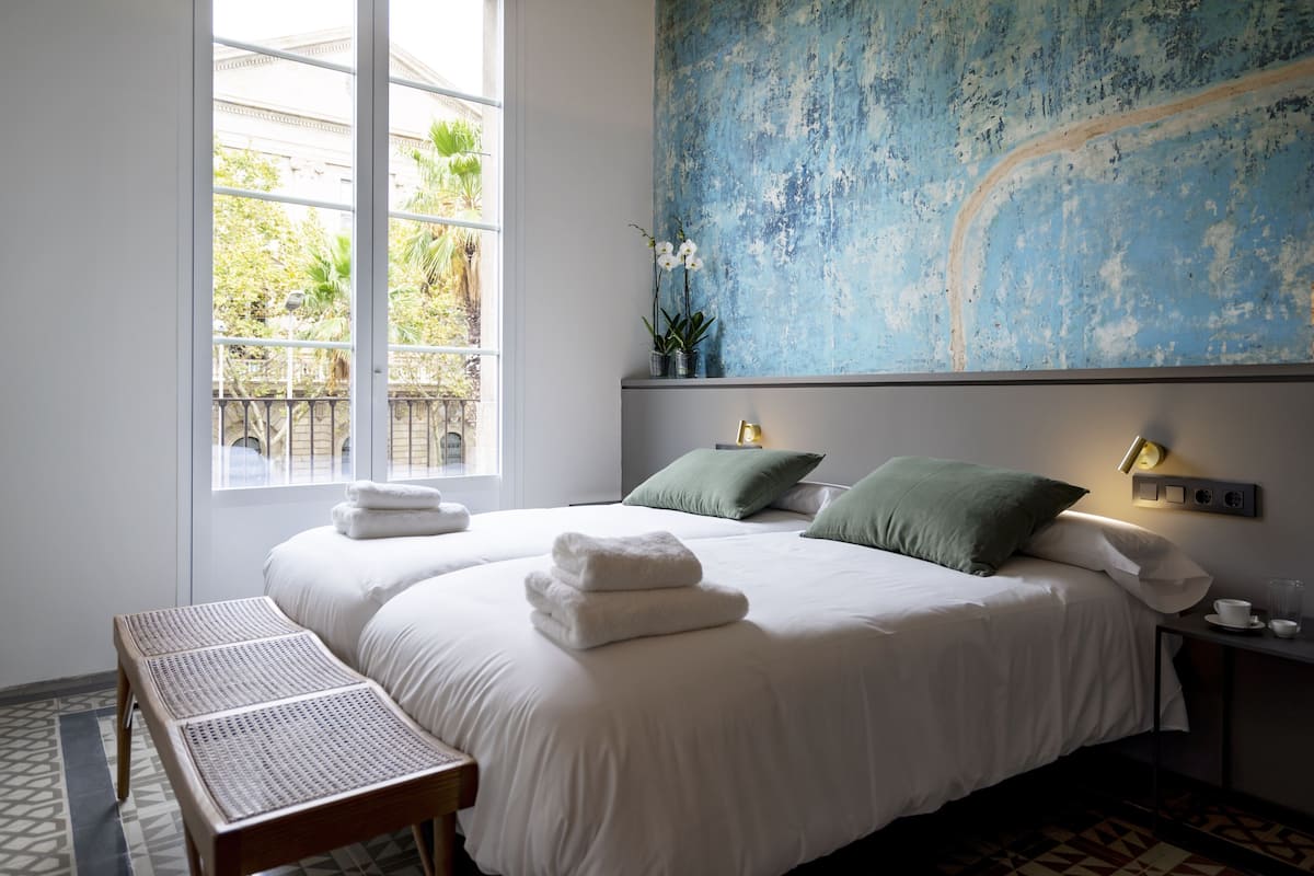 A neatly made bed with white linens and folded towels, a bedside table with a cup and a small plant, and a large window with a view of greenery outside.