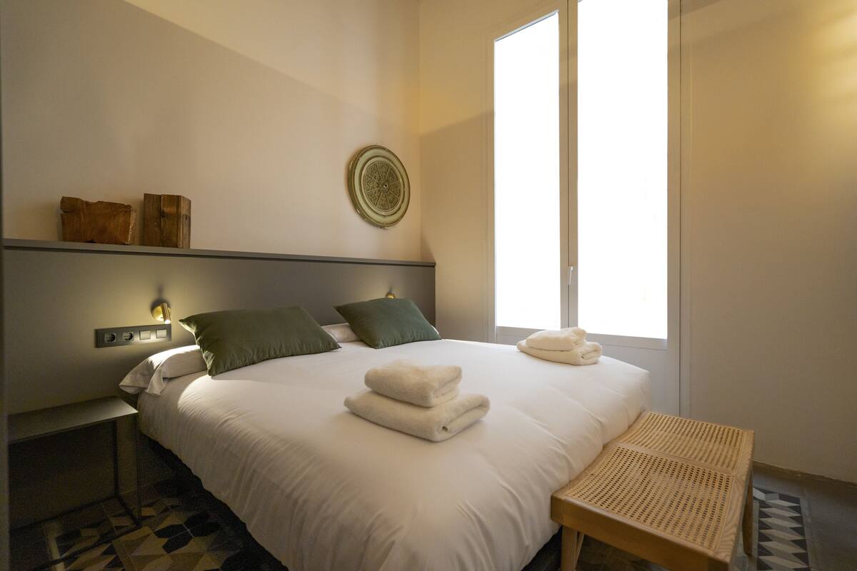 A neatly made bed with two pillows, a bedside table, a wall-mounted light, and a window with blinds.