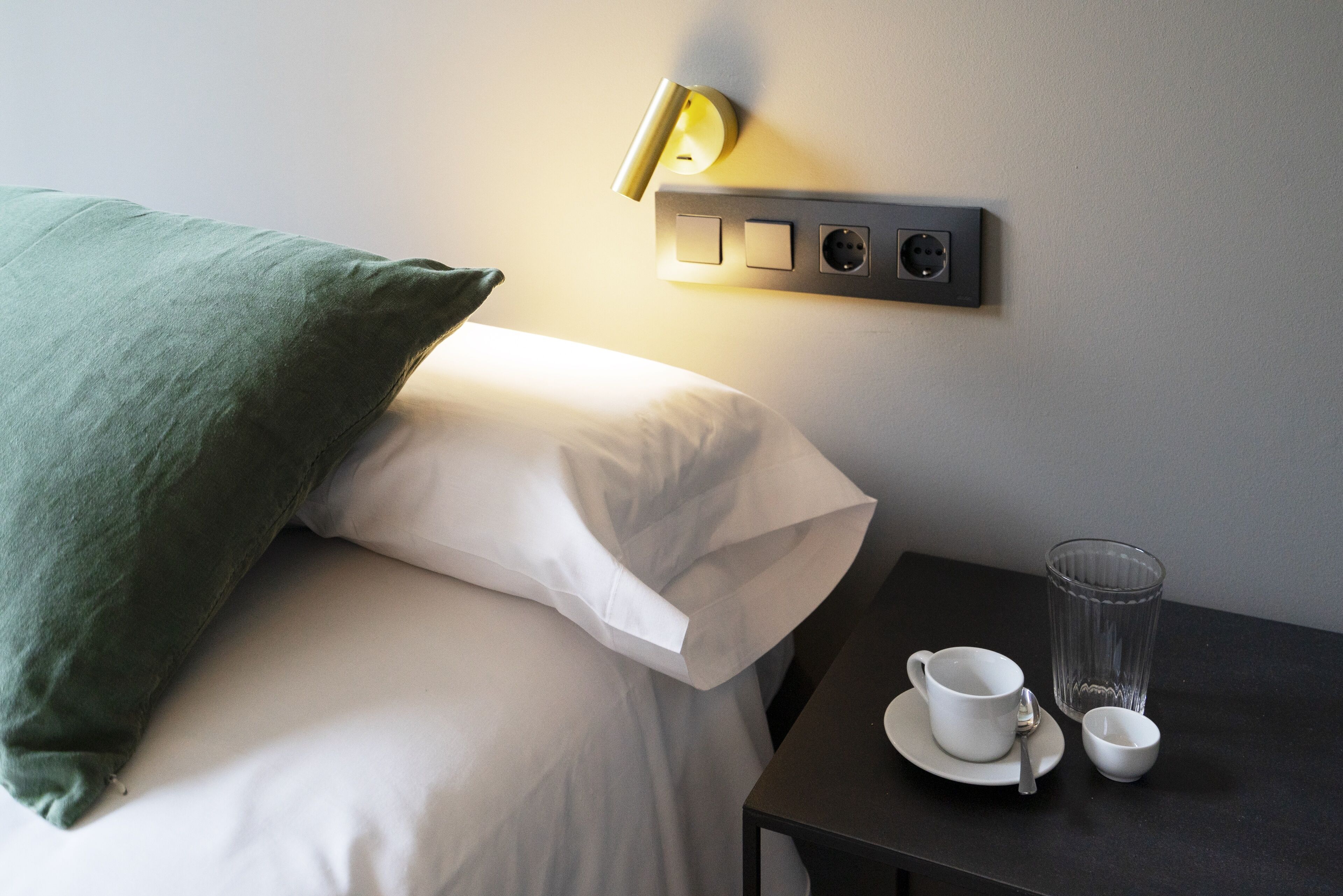 Una cama bien hecha con un almohadón verde y ropa blanca, una mesita de noche con una taza, un vaso y una cuchara, y un aplique de pared.