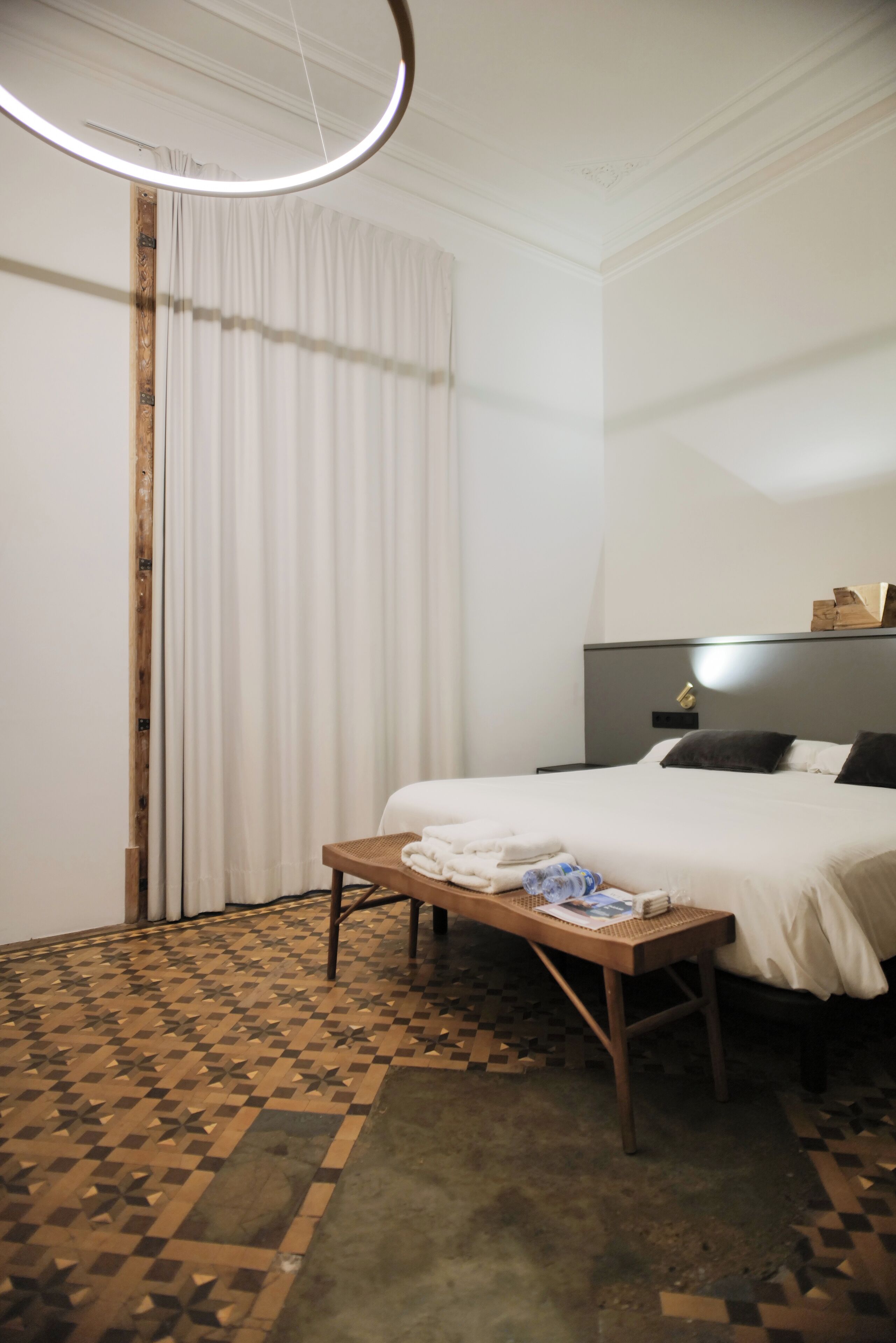 Habitación de hotel con cama, mesita de noche con toallas, cabecero con luz y suelo estampado.