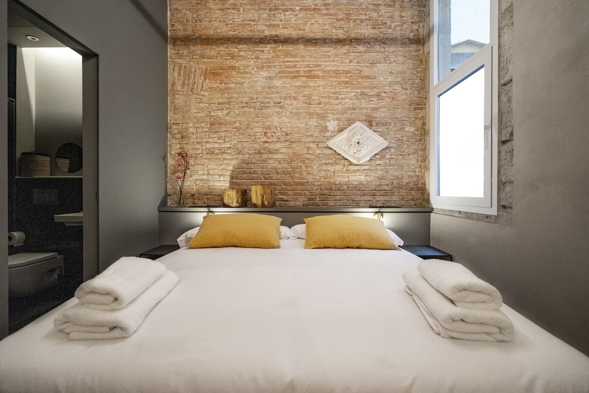 A hotel room with a brick accent wall, a bed with white linens and two yellow pillows, and a window on the right side.