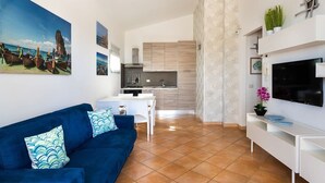 A modern living room with a blue sofa, a TV, and a kitchen in the background. - Corrado Residence (Blu) - Apartment near Marzamemi (Reitani)