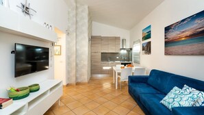 A modern living room with a flat-screen TV, a blue sofa, and a dining area with a white table and chairs. - Corrado Residence (Alba) -Apartment near Marzamemi (Reitani)