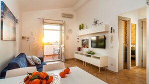 A modern living room with a sofa, a dining table with oranges, a TV, and a sliding glass door leading to a balcony. - Corrado Residence (Alba) -Apartment near Marzamemi (Reitani)