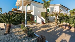 A modern two-story house with a white exterior, a covered outdoor area, and a well-maintained garden with various plants and a palm tree. - Corrado Residence (Alba) -Apartment near Marzamemi (Reitani)