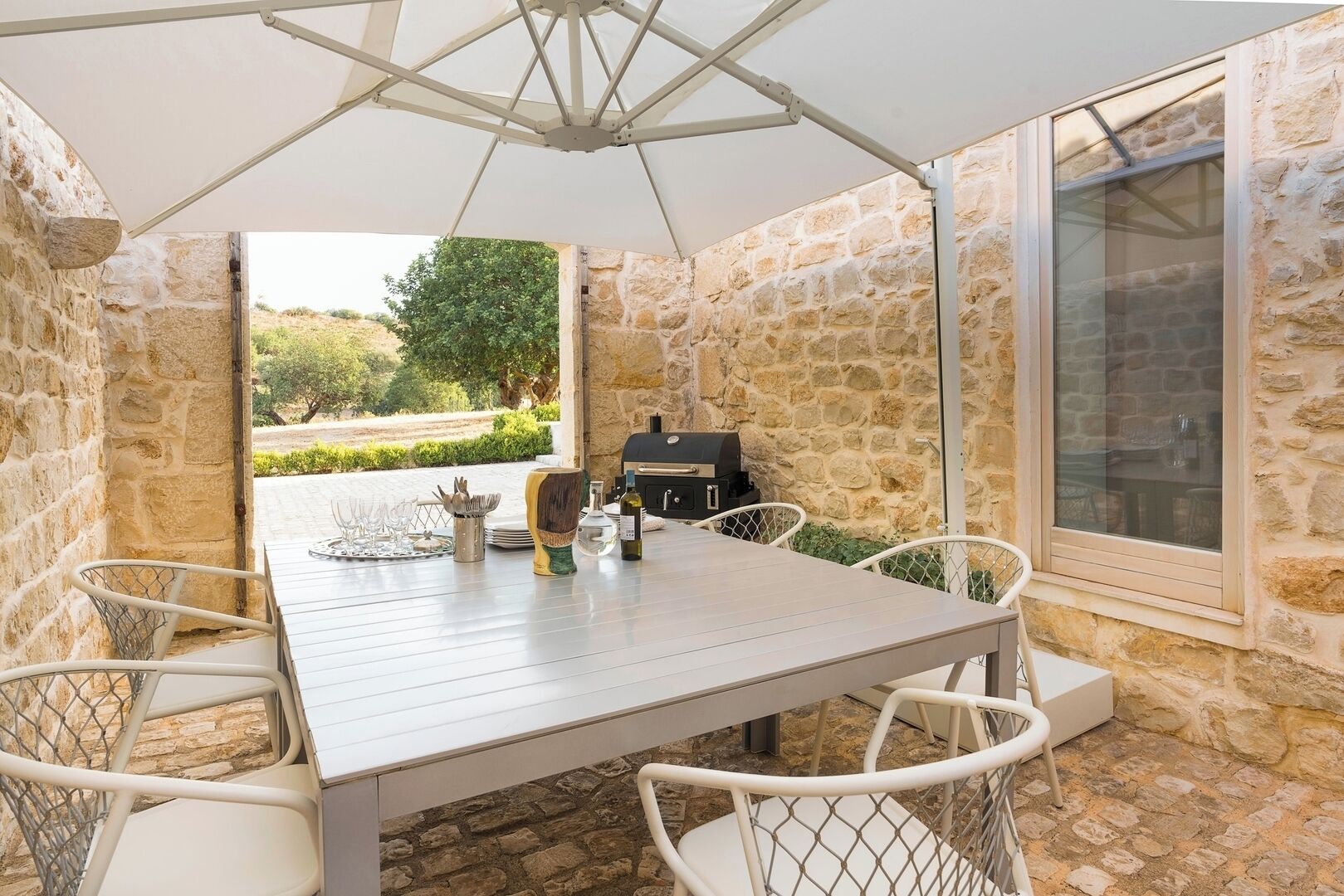 Patio esterno con muro in pietra, tavolo con sedie, barbecue e vista sul paesaggio.