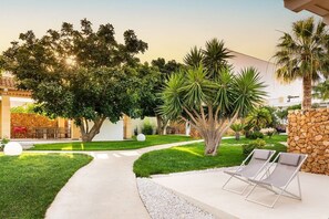 Área externa bem cuidada, com calçada pavimentada, cadeiras de descanso e diversas árvores e plantas.