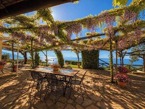 An outdoor patio with a table and chairs, surrounded by greenery and a view of the sea.