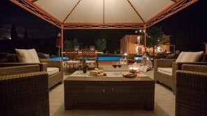 Een terras met rieten meubilair, een centrale tafel met drankjes en snacks, en een zwembad op de achtergrond.