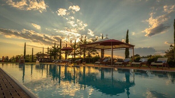 Ein Swimmingpool mit Liegestühlen und Sonnenschirmen, umgeben von Bäumen und einem Sonnenuntergangshimmel.