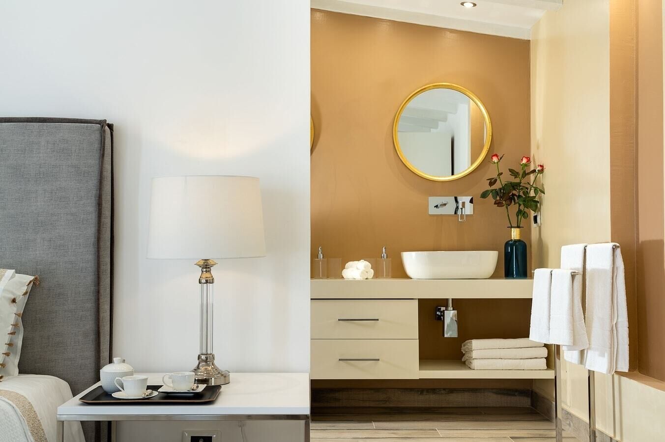 Ein modernes Hotelzimmer mit Bett, Nachttisch mit Lampe, Tablett mit Tee-Service, Badezimmer mit Waschbecken, Spiegel, Handtuchhalter und einer Vase mit Blumen.