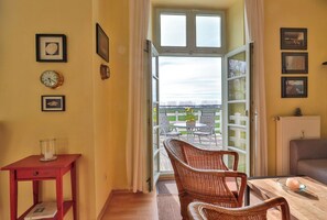 Una habitación con una silla de mimbre, una mesita, una mesita auxiliar roja y vistas a un área exterior con una mesa y sillas a través de una puerta de cristal.