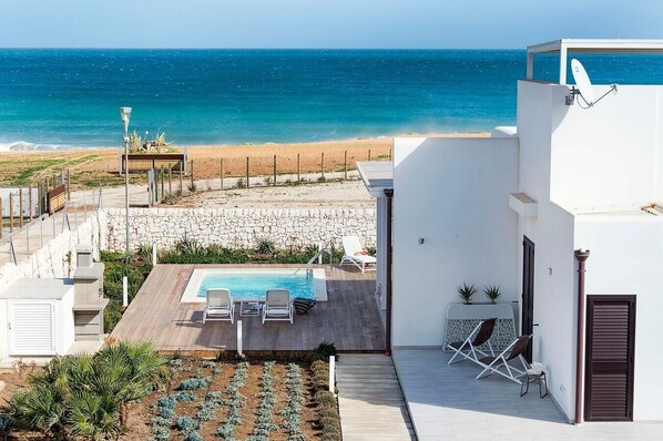 A pool area with lounge chairs, a view of the beach, and a stone wall.