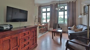 Ein Wohnzimmer mit Fernseher, Holzschrank, Esstisch, Stühlen, einer Couch und großen Fenstern.