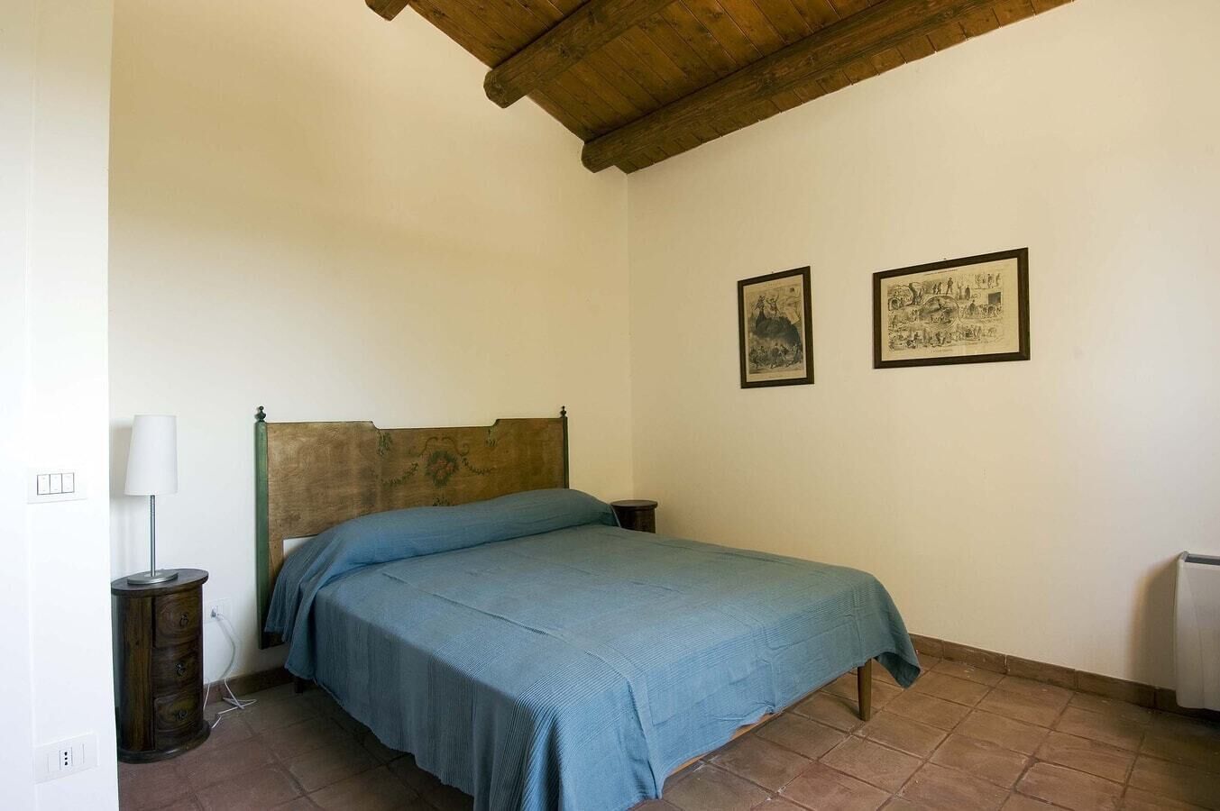 A bedroom with a wooden headboard, a single bed with a blue bedspread, two framed pictures on the wall, a bedside lamp, and a small wooden nightstand.