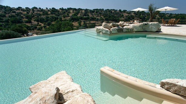 A clear swimming pool with a stone edge, surrounded by lounge chairs and umbrellas, with a view of green hills in the background.