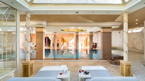A luxurious spa area with a large pool, two lounge chairs, and a reception area visible through glass doors.