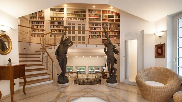 A spacious living area with a high ceiling, a staircase, and a large bookcase filled with books.