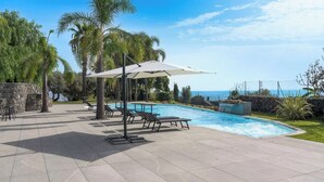 Un espace piscine avec des chaises longues et des parasols, un mur en pierre et une vue sur l’océan.