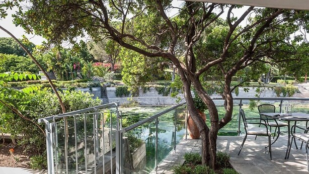 Un espace extérieur paisible, doté d’un pont de verre, d’un coin salon avec tables et chaises, et d’une végétation luxuriante.