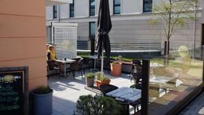 An outdoor cafe area with tables and chairs, potted plants, and a glass wall with a reflection. - Der Zweckmäßig Ausgestattete Bungalow in Börgerende mit ca. 42 qm Wohnfläche Verfügt Über Eine gut Abgeschirmte Sonnenterrasse in Süd-/westlage (Börgerende)