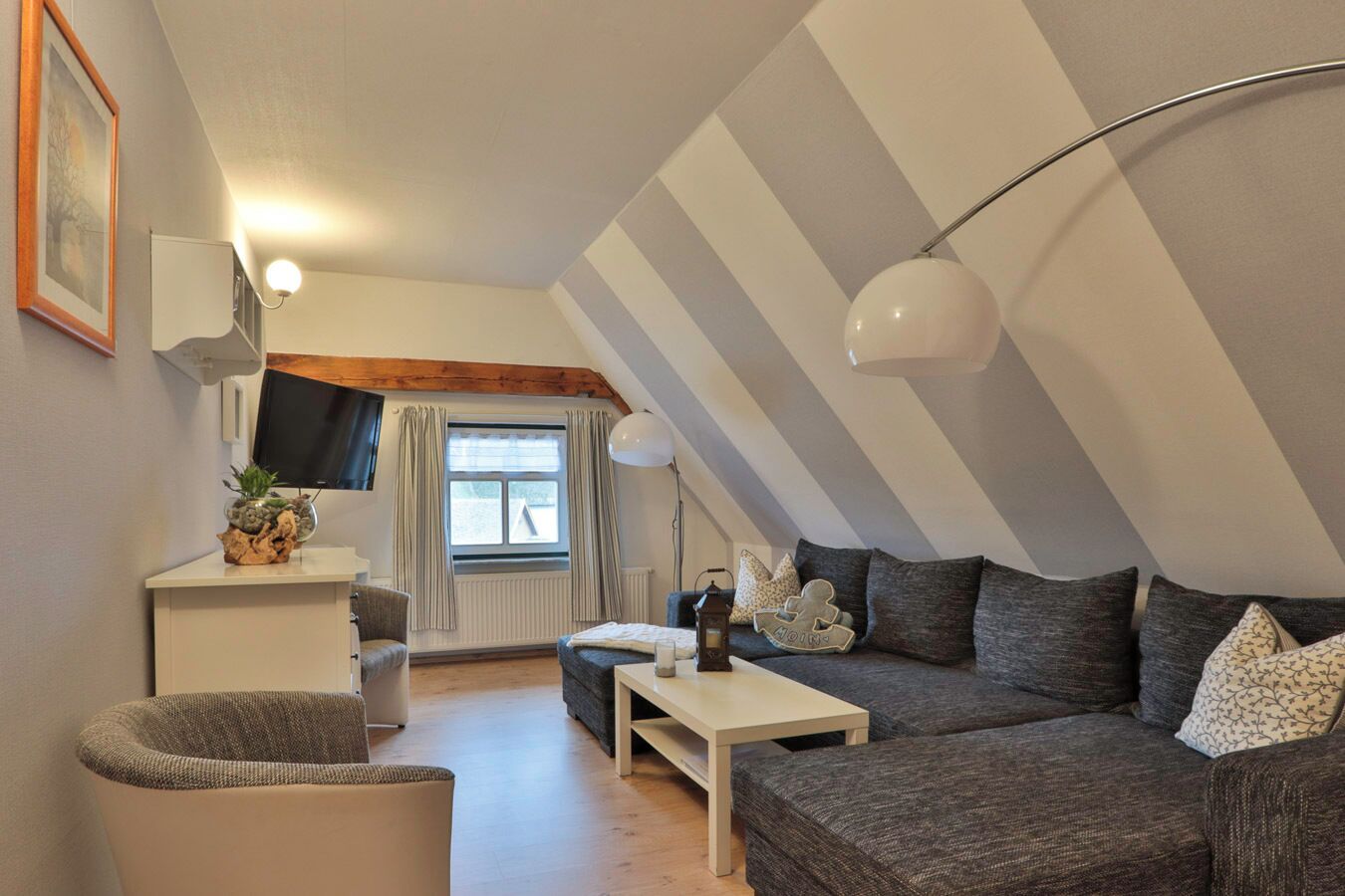 Ein Loft-Zimmer mit schrägem Dach, einer Sofagarnitur, einem kleinen Tisch, einem Sessel, einem Fernseher und einem Fenster mit Vorhängen.