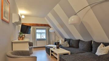 Ein Loft-Zimmer mit schrägem Dach, einer Sofagarnitur, einem kleinen Tisch, einem Sessel, einem Fernseher und einem Fenster mit Vorhängen.