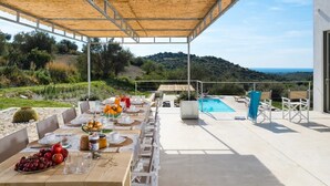 Un espace extérieur aménagé pour les repas, offrant une vue sur la mer et les collines, ainsi qu’une piscine et un coin salon abrité.