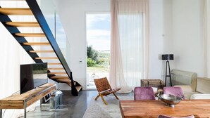Un salon moderne avec un escalier en bois, un téléviseur à écran plat, un coin repas avec une table en bois et une vue sur l’extérieur grâce à de grandes fenêtres.