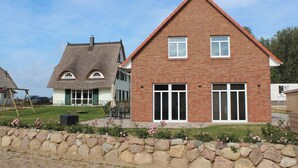 Une maison en briques avec une grande fenêtre et une porte, un mur de pierres à l’avant et une cour gazonnée.