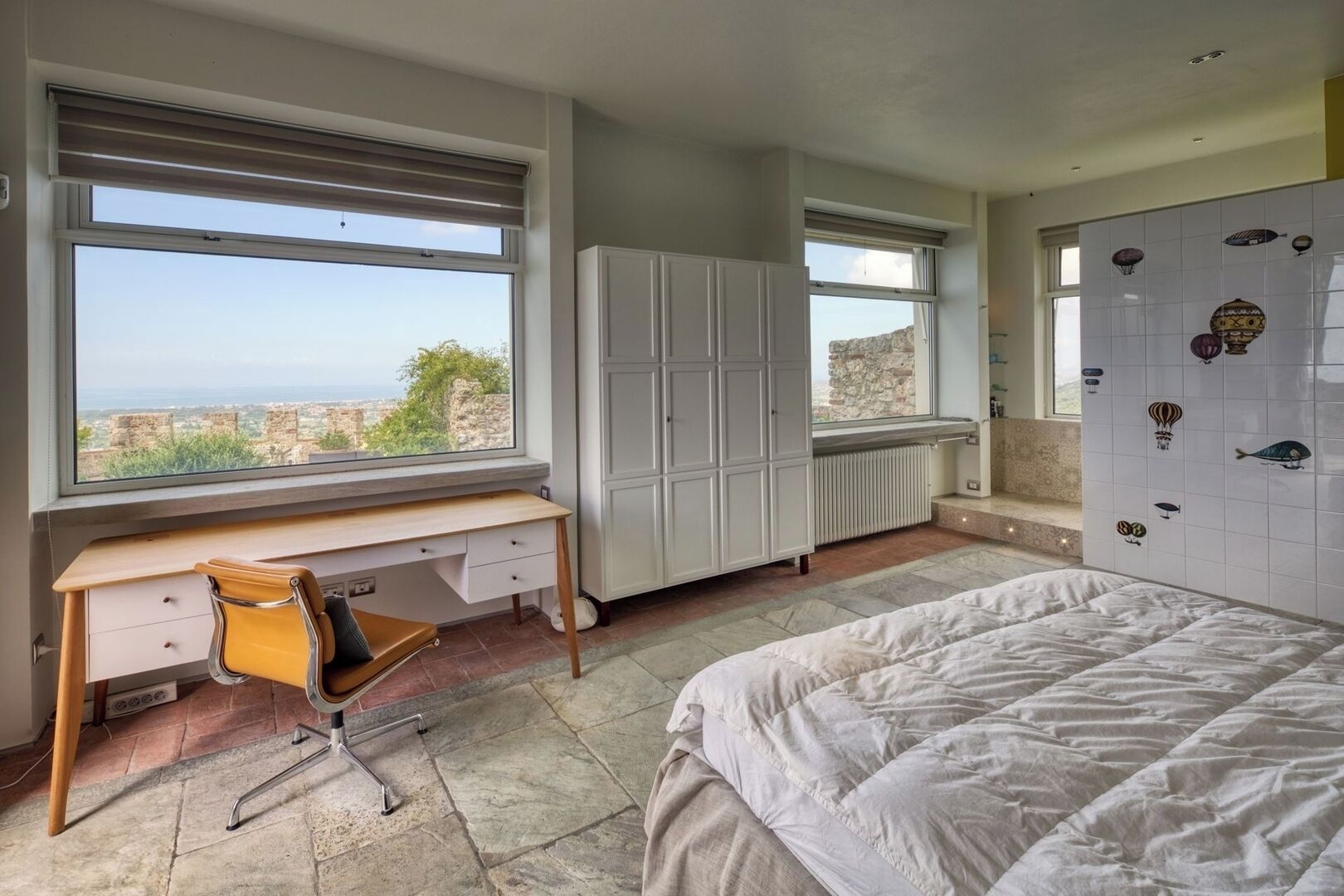 A bedroom with a bed, a desk, and a view of the city through large windows.