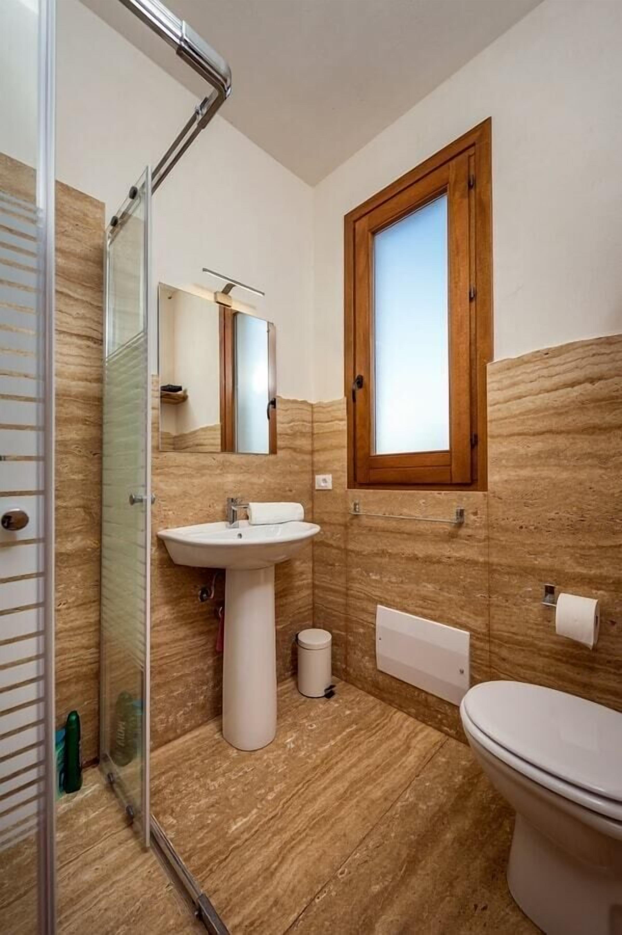 Ein Badezimmer mit Duschkabine aus Glas, einem Standwaschbecken, einer Toilette und einem Fenster mit Holzrahmen.
