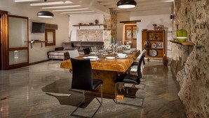 Een eetgedeelte met een houten tafel voor zes personen, omringd door zwarte stoelen. Achtergrond is een stenen muur en een houten kast met servies.