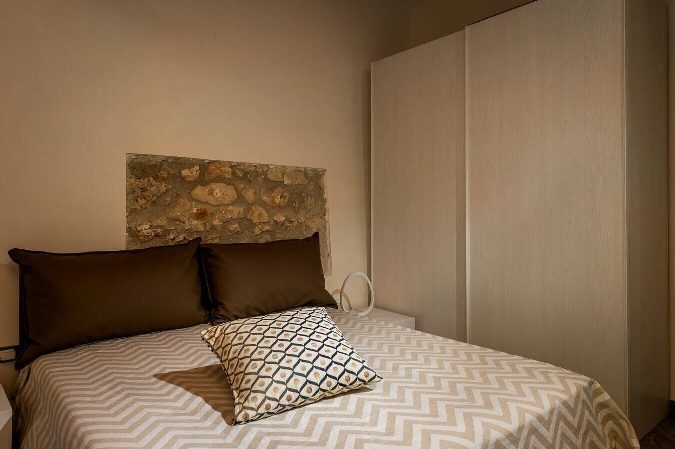 Una cama con ropa de cama estampada, dos almohadas, una luz de aro y una pared de piedra detrás de la cama.