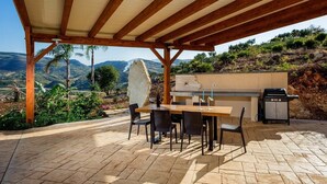 Een terras buiten met een houten pergola, een tafel met stoelen, een barbecue en uitzicht op de heuvels.