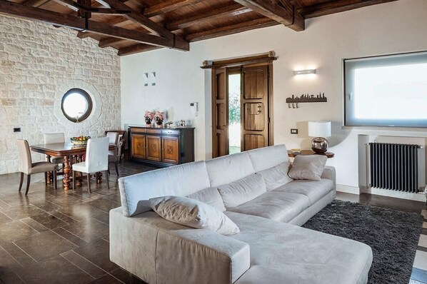 A living room with a large beige sofa, a dining table with chairs, a stone wall, a wooden cabinet, and a radiator.