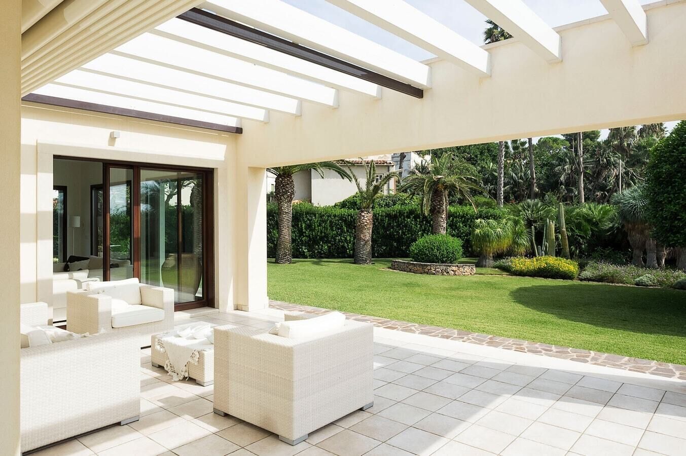 Ein moderner Außenbereich mit einer weißen Sofagarnitur und einem kleinen runden Tisch, einer Glastür, die auf eine Terrasse führt, und einem gepflegten Garten mit verschiedenen Pflanzen und einem Rasen.