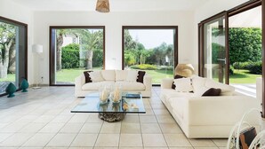 Ein geräumiges Wohnzimmer mit einem großen Glasfenster zum Garten, einem weißen Sofa, einem Glastisch und einer Pendelleuchte.