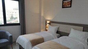 A hotel room with two beds, a gray armchair, and a view of buildings outside the window. - Hôtel du Lac Béjaïa (Béjaïa)