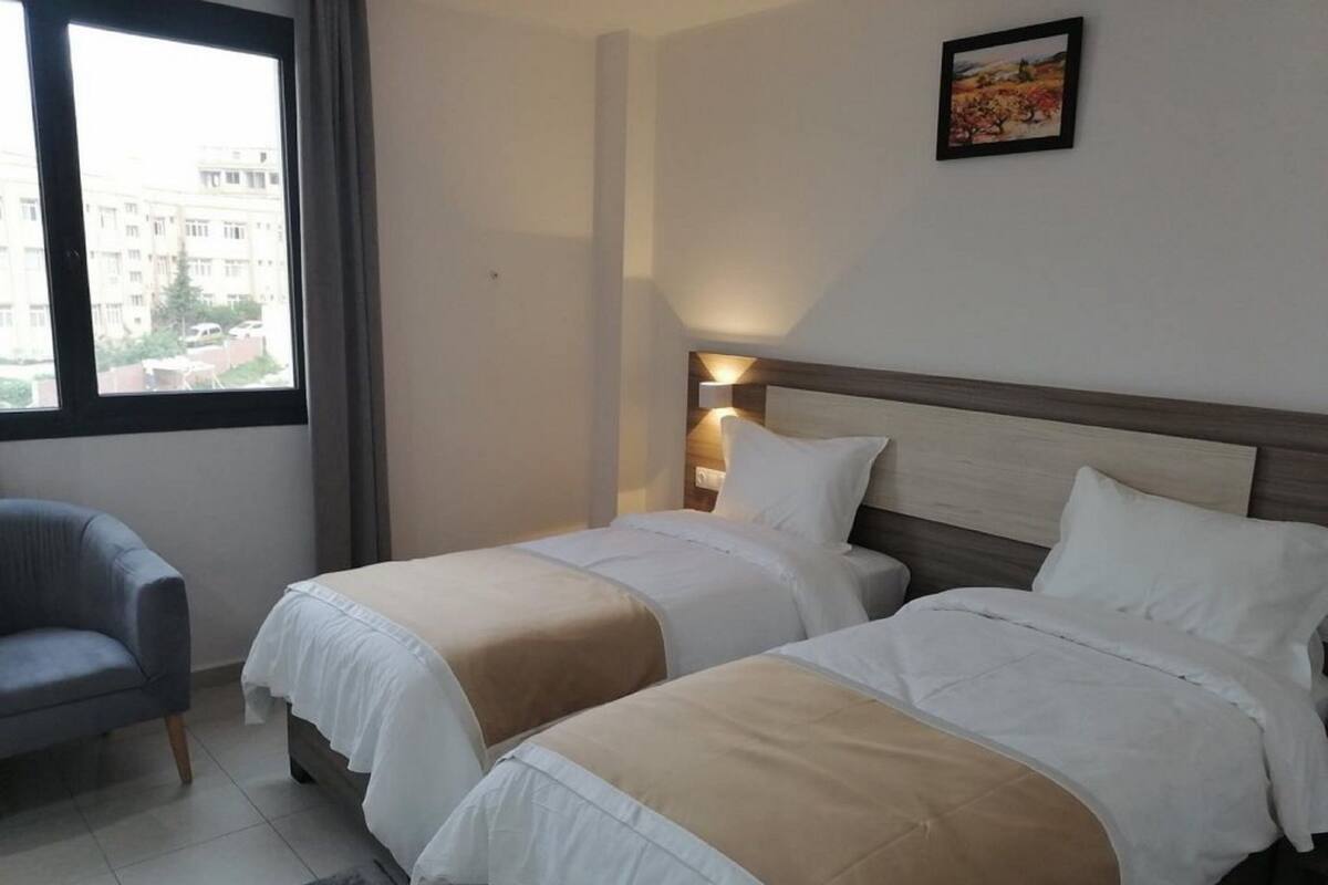 Ein Hotelzimmer mit zwei Betten, einem grauen Sessel und Blick auf Gebäude.