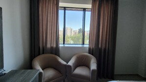 A hotel room with two armchairs, a large window with curtains, and a view of buildings and trees. - Hôtel du Lac Béjaïa (Béjaïa)